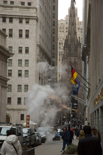 Tall buildings on Wall Street create a slender canyon; a church steeple is at the end of the street.