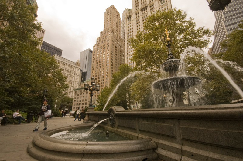 Jets of water stream towards a multi-tiered fountain in a plaza surrounded by skyscrapers.
