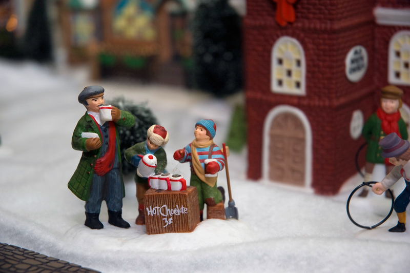 Figurines of a man drinking hot cocoa he's bought from two kids with a stand.